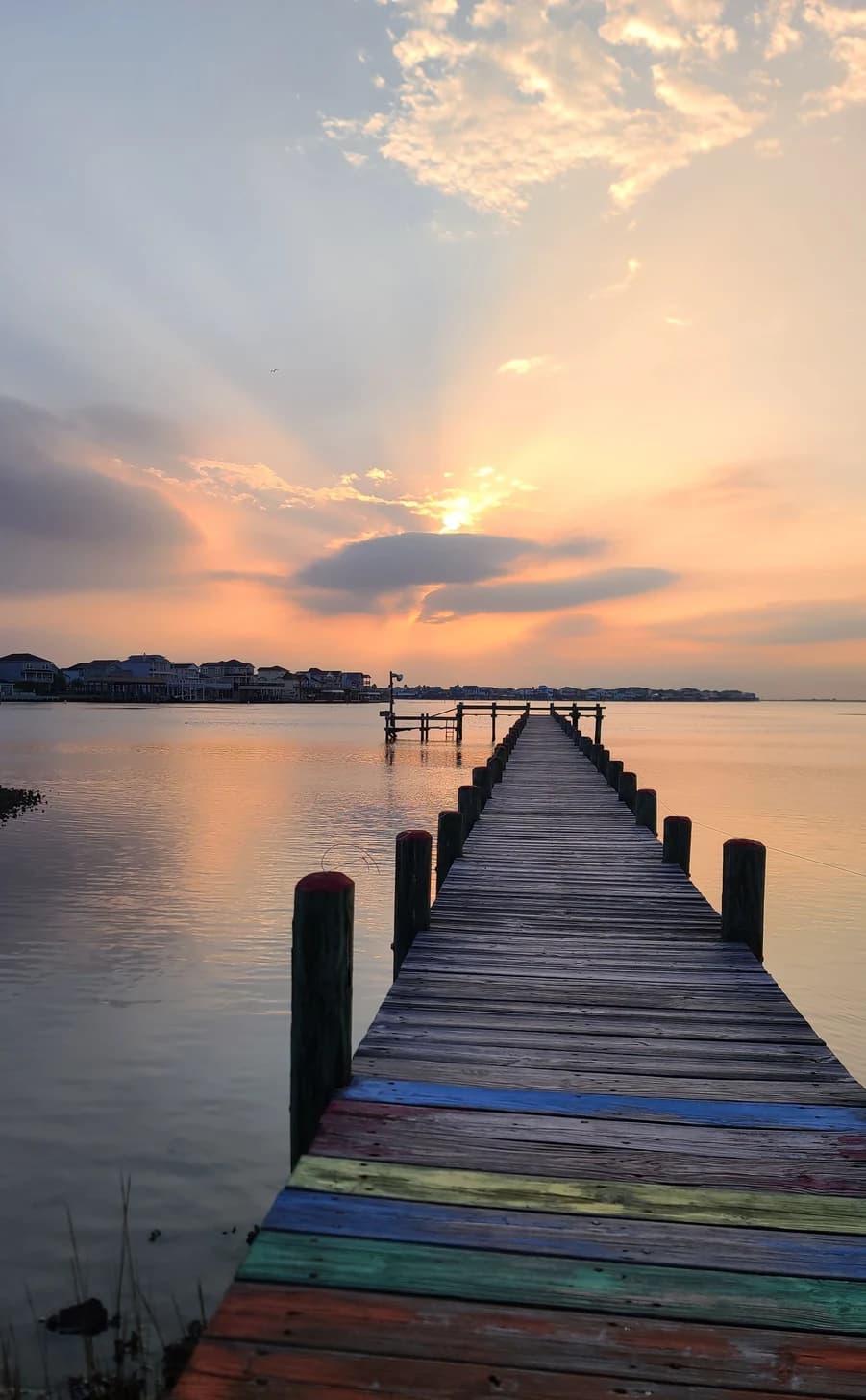 A long pier with weathered multicolor boards points toward calm water and a bright sky.
