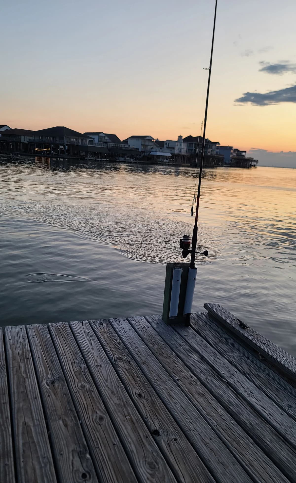 A fishing rod stands in a holder on a weathered wooden pier above calm evening water.