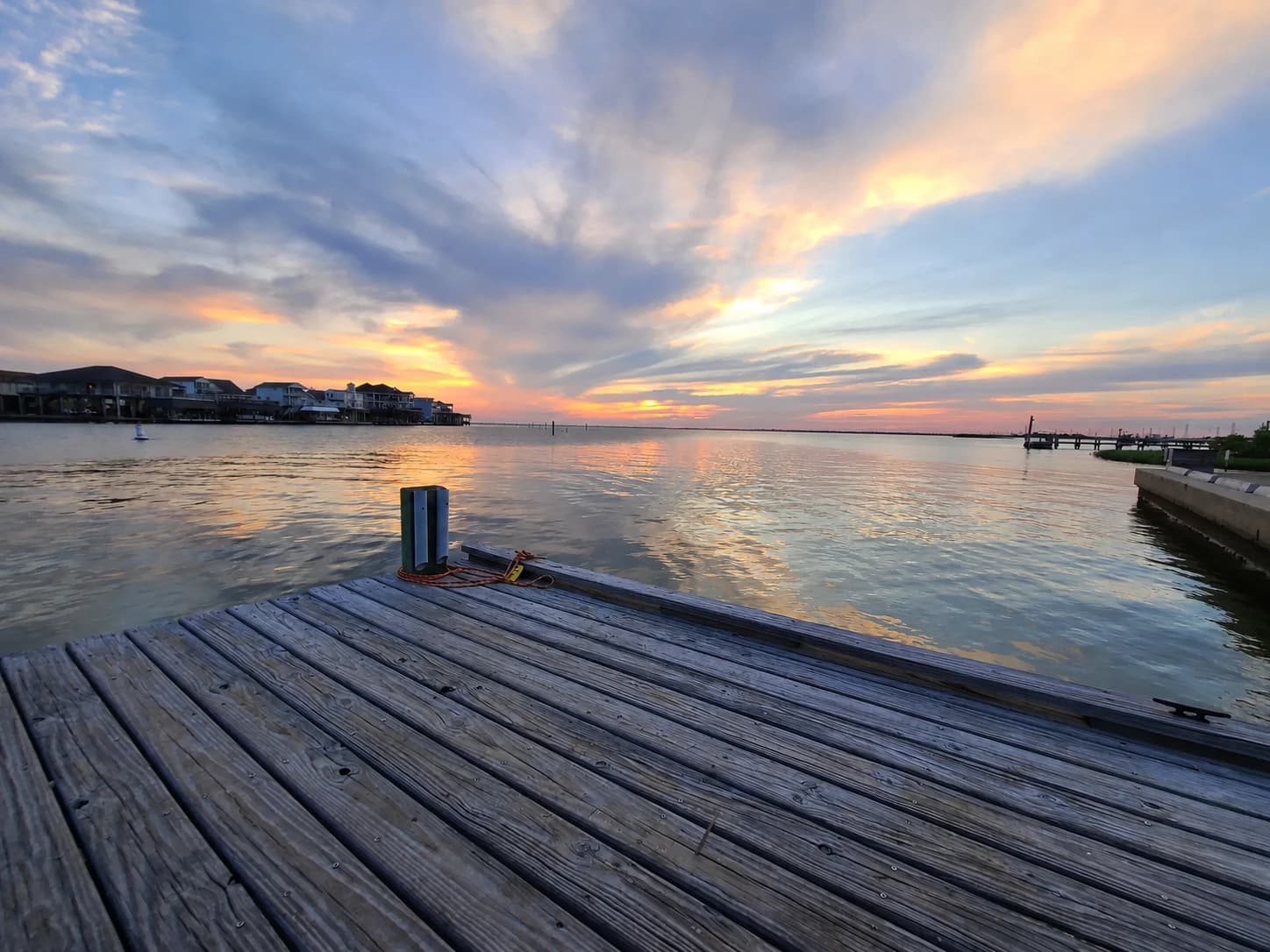 A wooden pier reaches into calm bay water beneath a pastel orange and blue sunset.