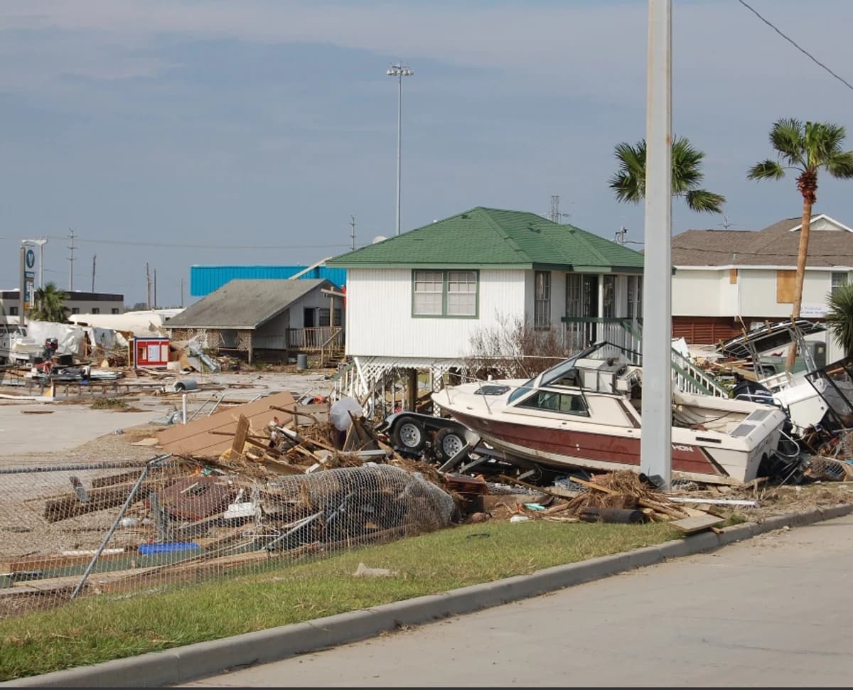 A historical photograph shows a boat, broken fencing, and storm debris around buildings after Hurricane Ike.
