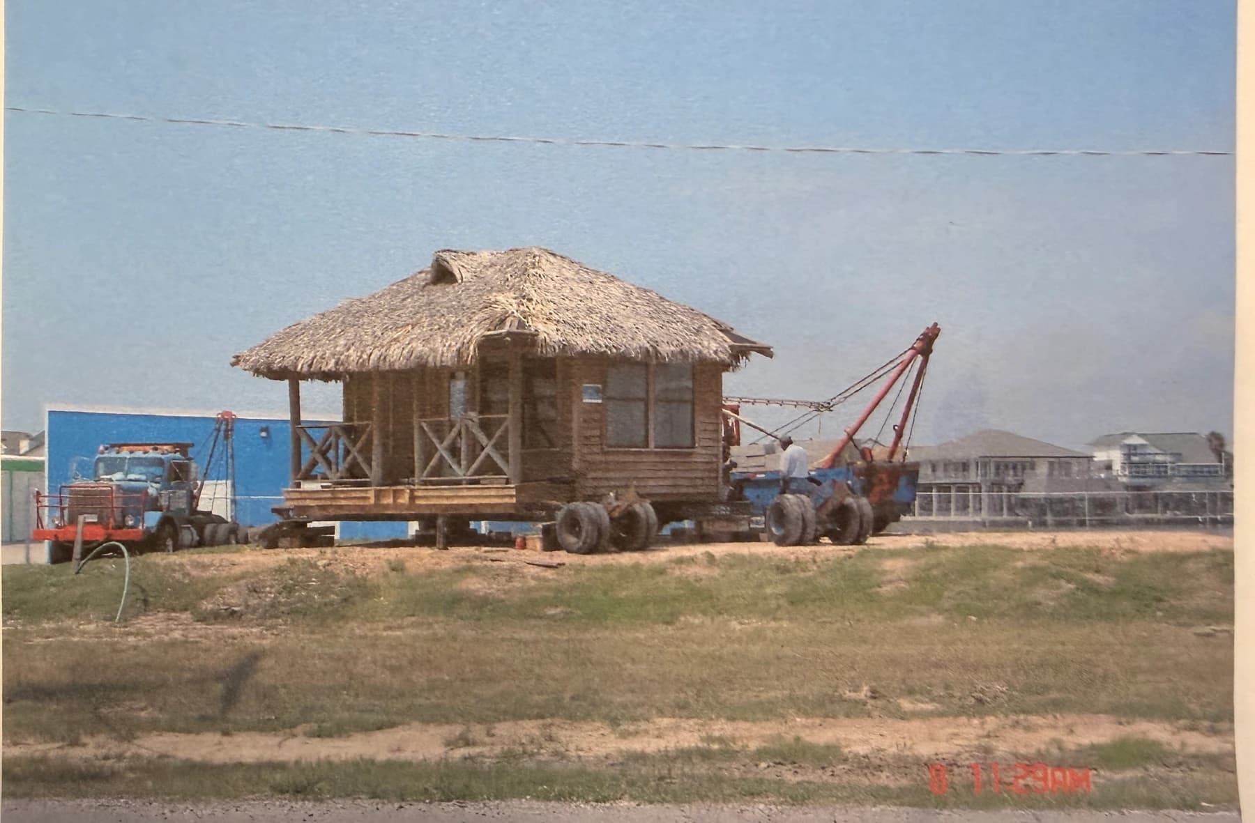 A historical photograph shows a small thatched-roof office building being moved by two tow trucks.