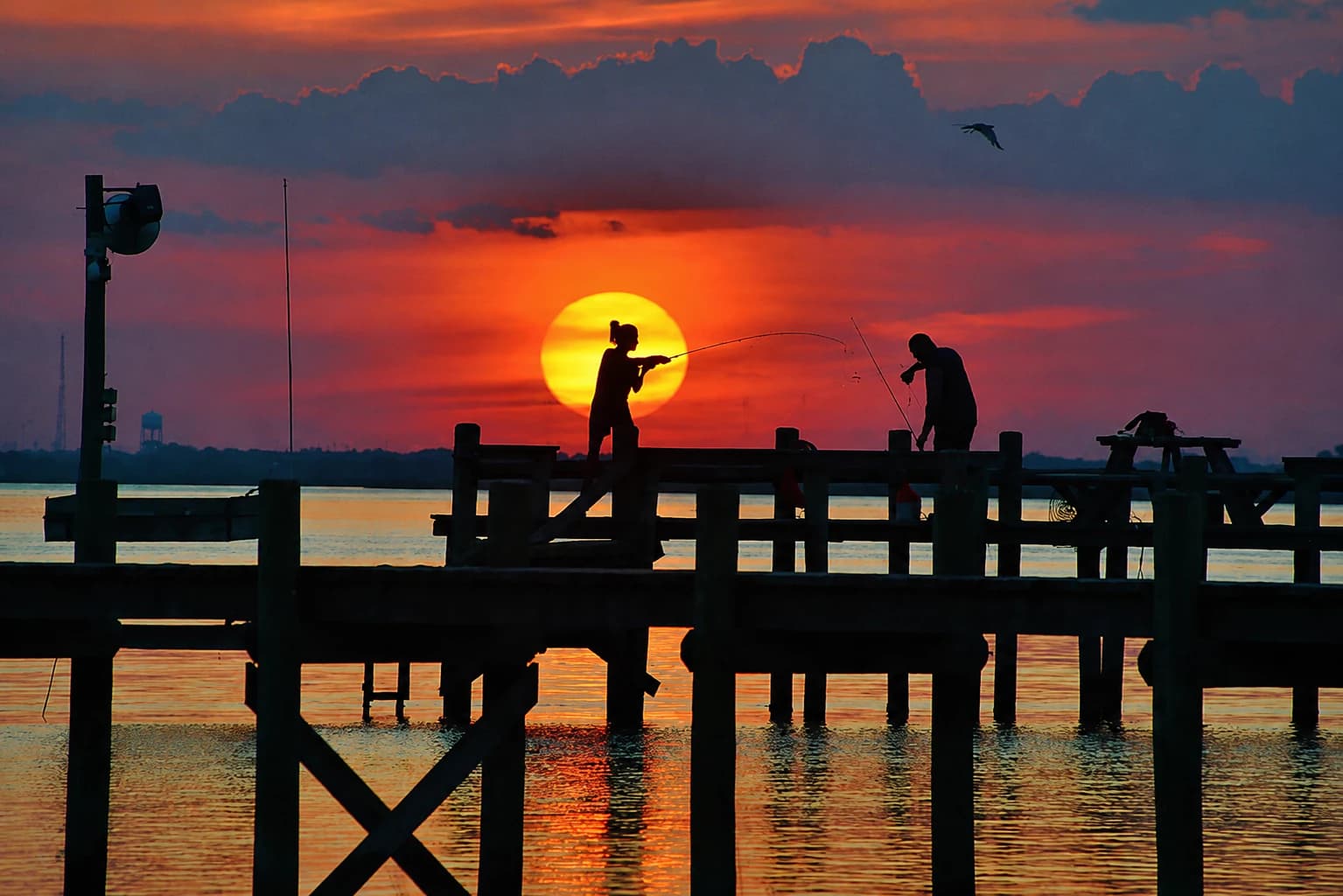 Two silhouetted people fish from a wooden pier in front of a large orange sunset over the bay.